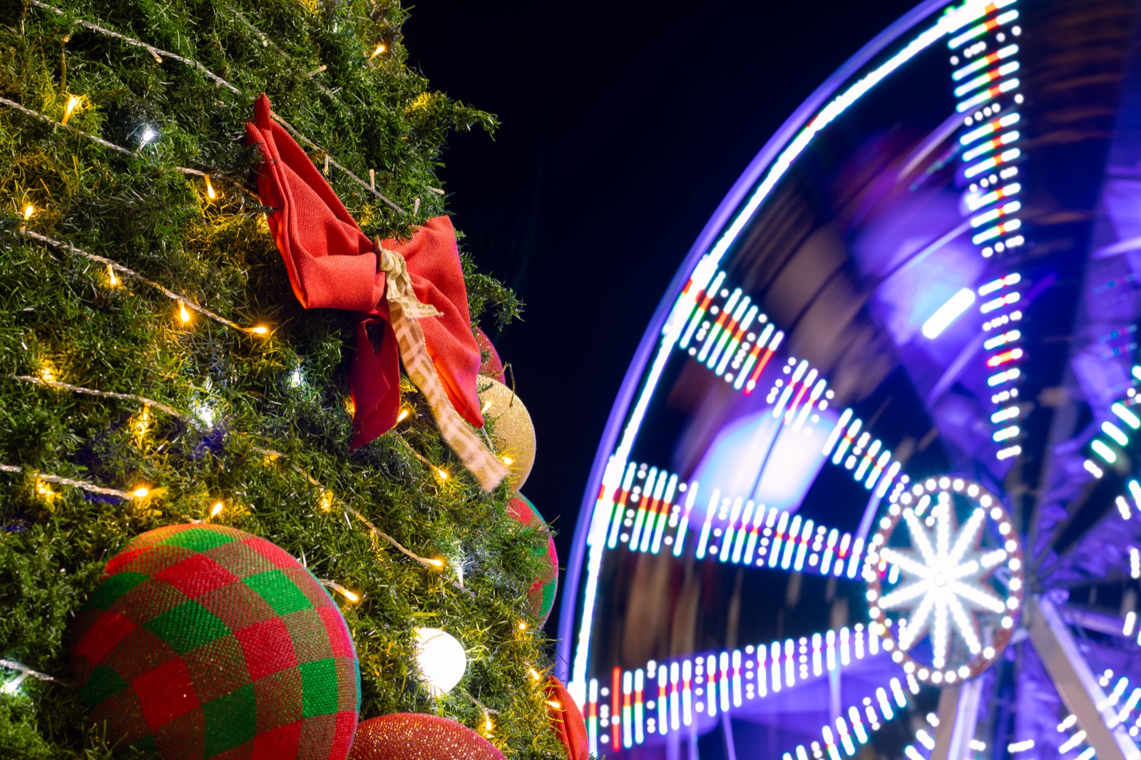 A decorated Christmas tree beside a ferris wheel at night, its lights blurred into streaks.