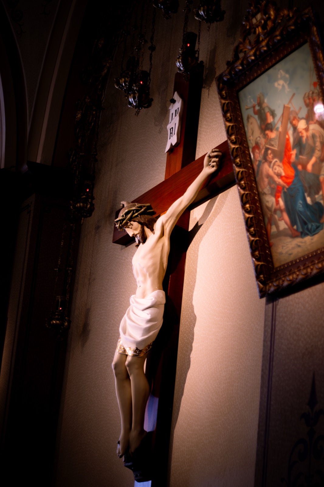 A carved crucifix lit dramatically on a church wall beside a framed painting.
