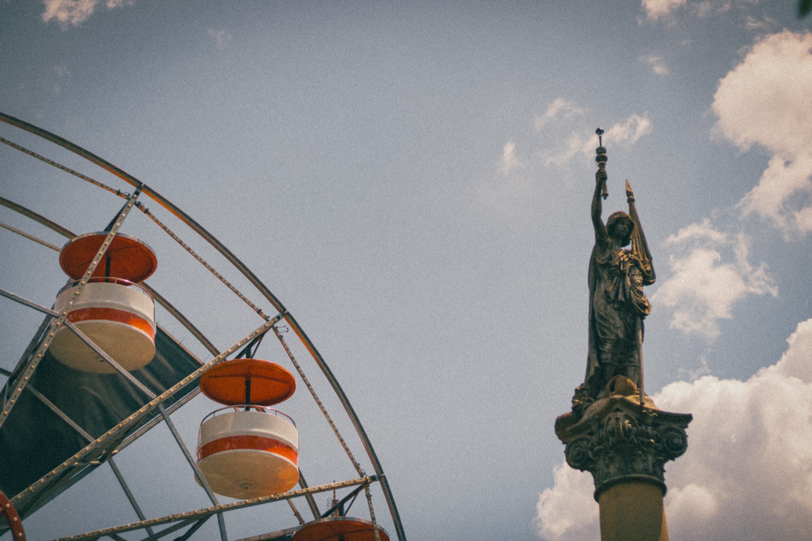 A vintage ferris wheel beside a monument statue under a cloudy sky.